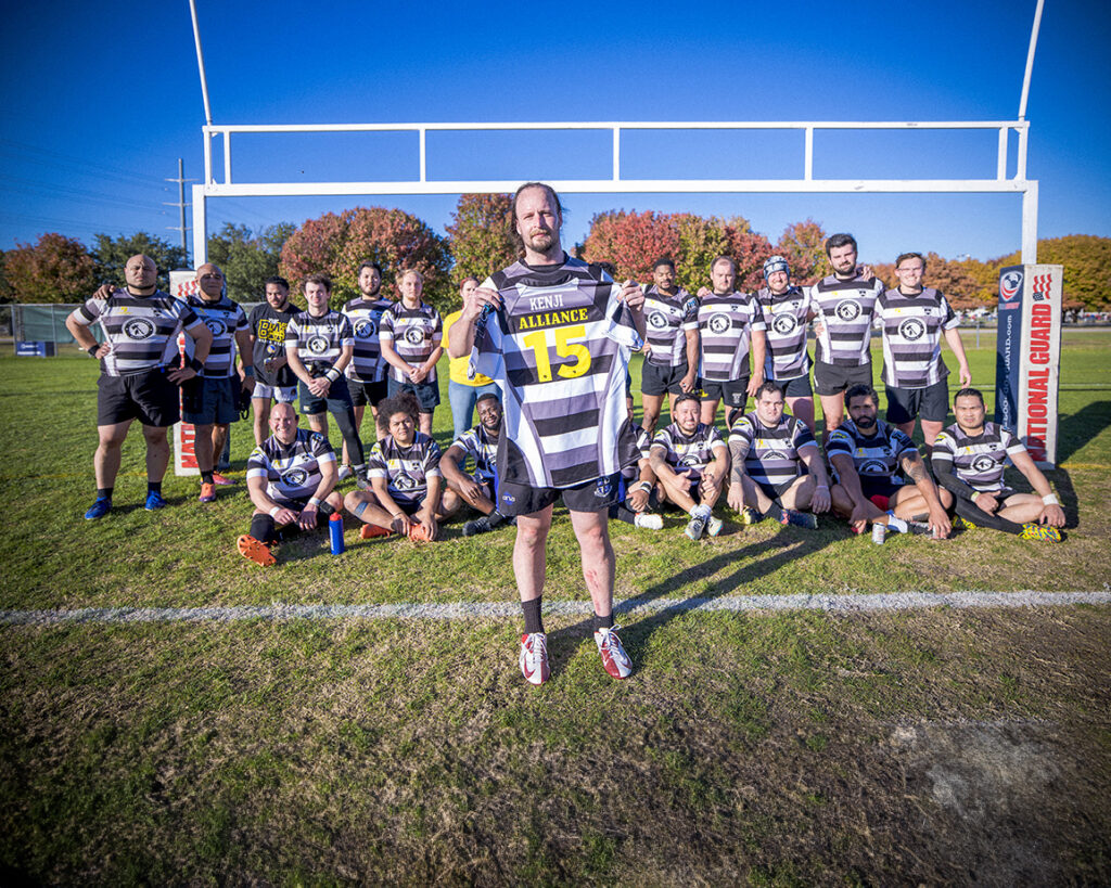 A rugby team posing in front of a goal post with one member holding up the jersey of a player that had passed.