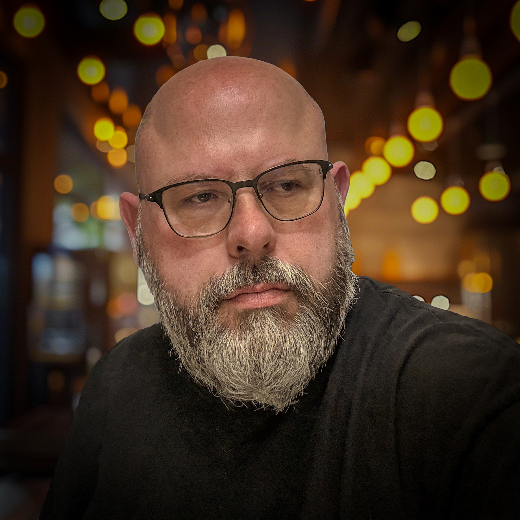 A caucasian male with a beard wearing glasses looking to his left in front of a backdrop with colored lights.