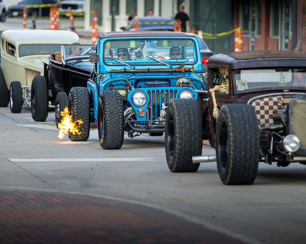 A blue lowered Jeep on a city street with flame coming from the exhaust amongst other hot rods