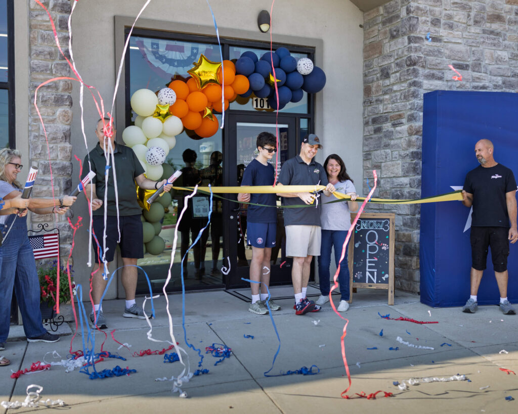 Three people stand in front of the entrance of a store. One of them is cutting a ribbon as part of a grand opening ceremony.
