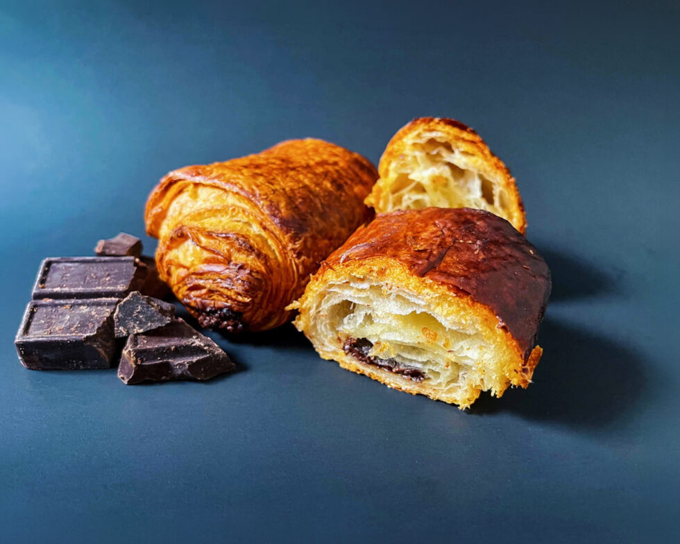 two chocolate croissants on a dark blue background. One is split open and there are pieces of chocolate against the other.