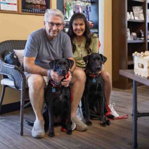 PetWants-Business-Photography-Texas-0011 A man and a woman sitting on a bench with two black dogs in front of them.